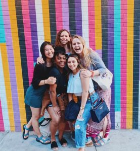 Five smiling BOLD Cohort 1 Scholars pose in front of a brick wall painted with vertical stripes in vibrant colors.